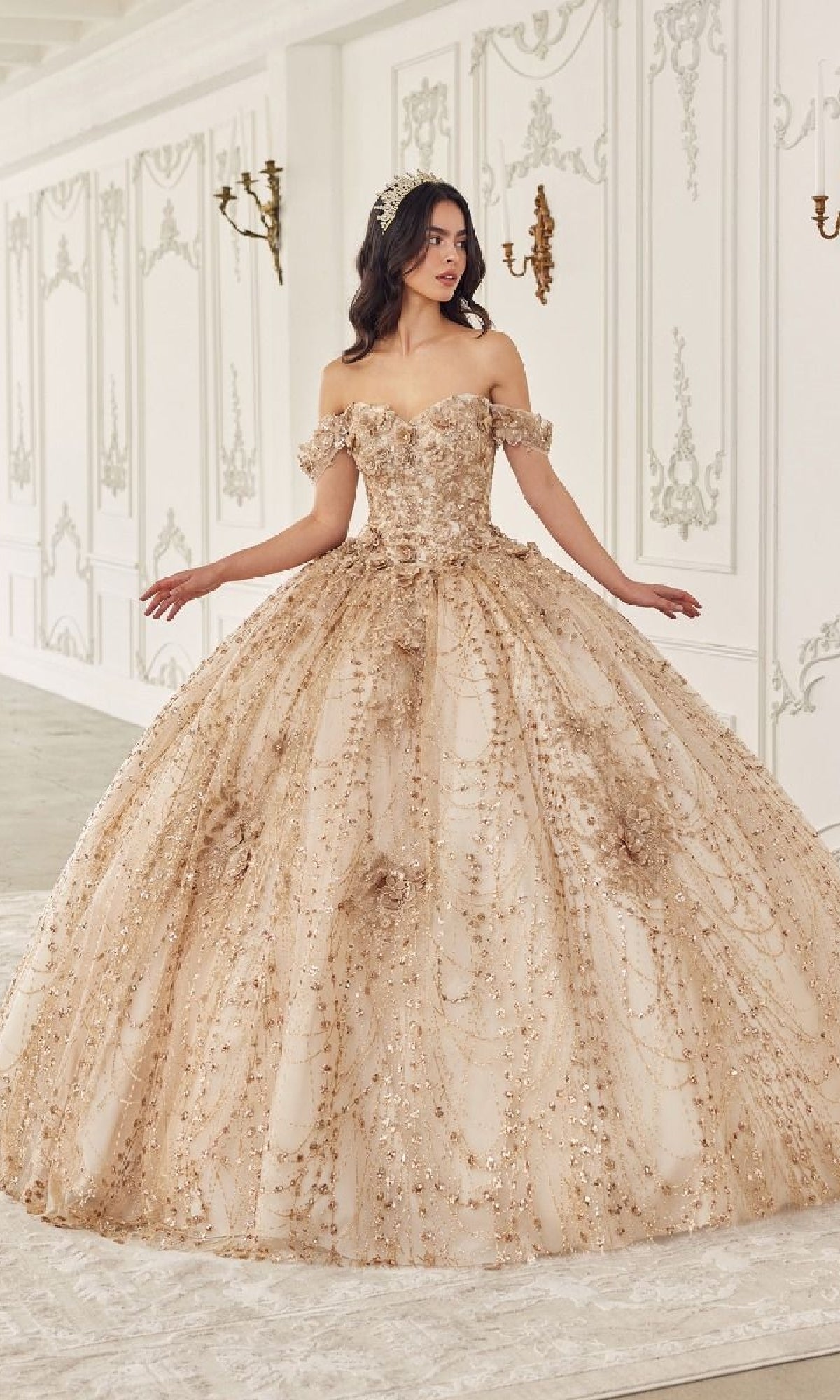 A woman stands in a decorated white room wearing the ornate, off-the-shoulder Quinceanera Dress 15712 by Ladivine, featuring a gold hue and full skirt, surrounded by wall moldings and sconces.